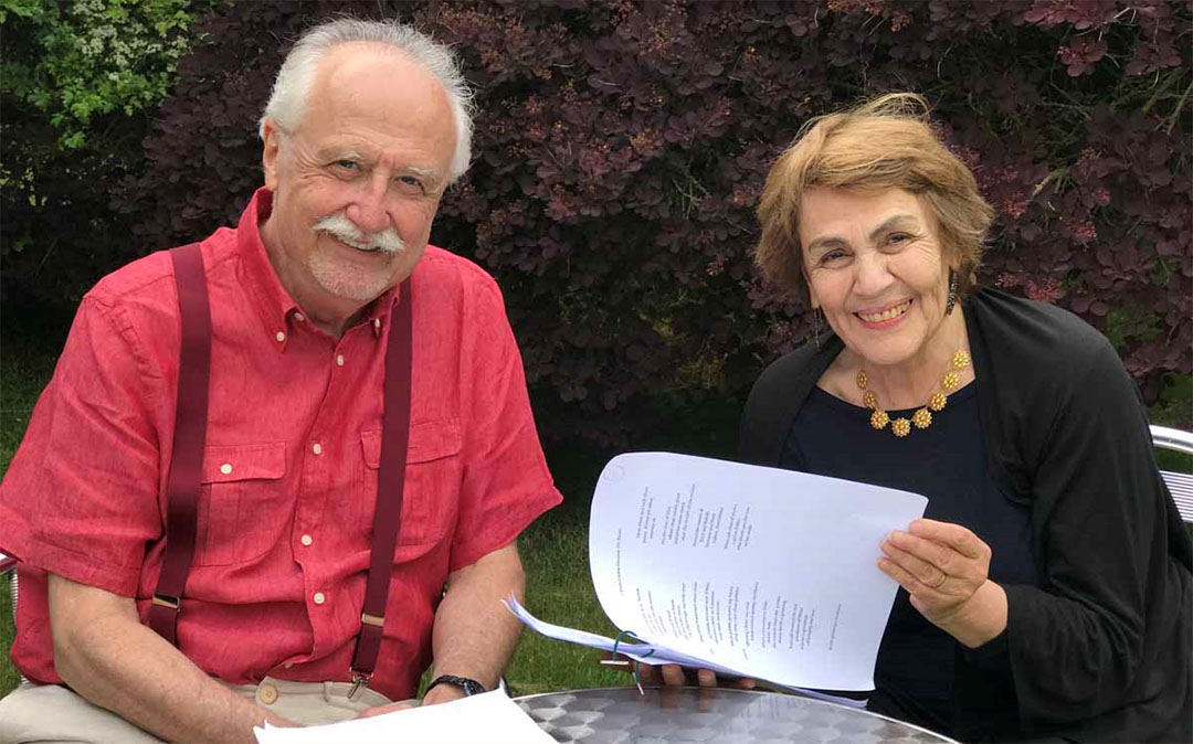 Michael Sells and Simone Fattal contributor photo, sitting at a metal table outside reading from a printer paper packet in front of hedge