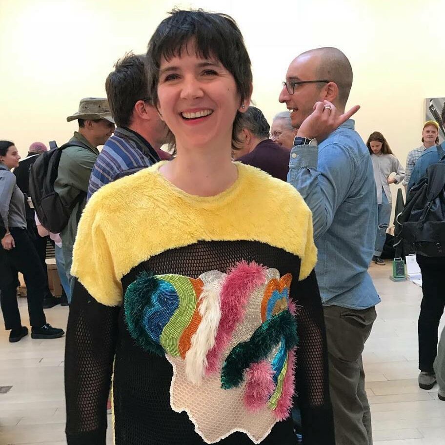 Rachael Wilson author photo, indoors with a mingling crowd behind her, smiling and wearing a bright multicolored, multitextured sweater