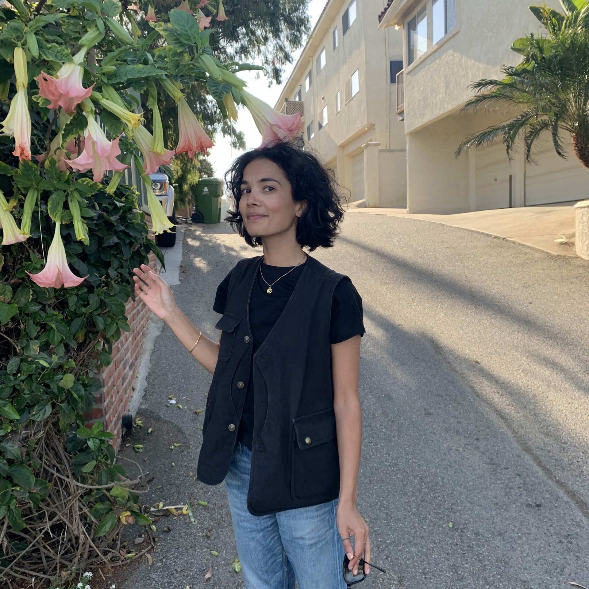 Alisha stands near a flowering tree on a sunlit street in California wearing a black shirt and jeans holding sunglasses in her hands