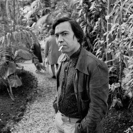 Mohammed Khair-Eddine stands on a path surrounded by tall tropical plants wearing a suit jacket and smoking a cigarette