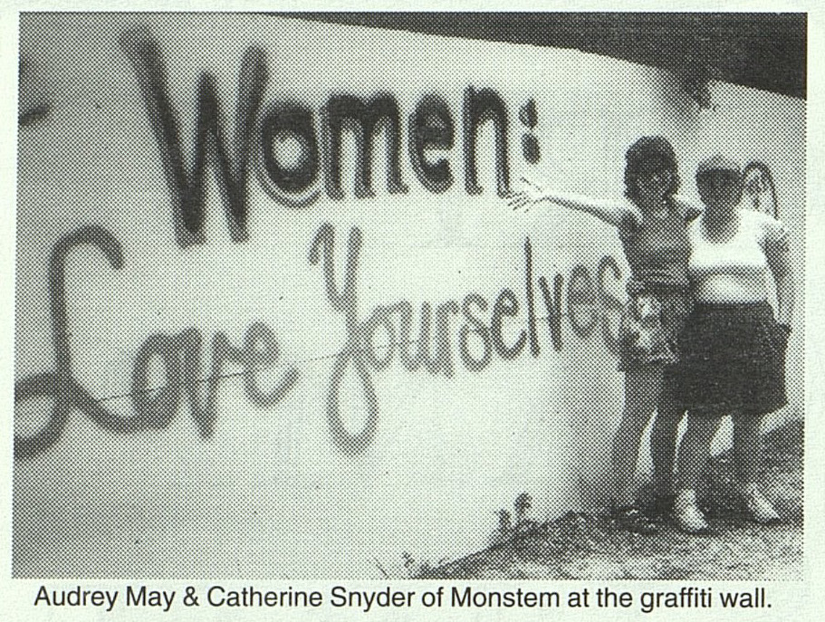 photograph of two women standing in front of a wall with graffiti that reads women love yourselves