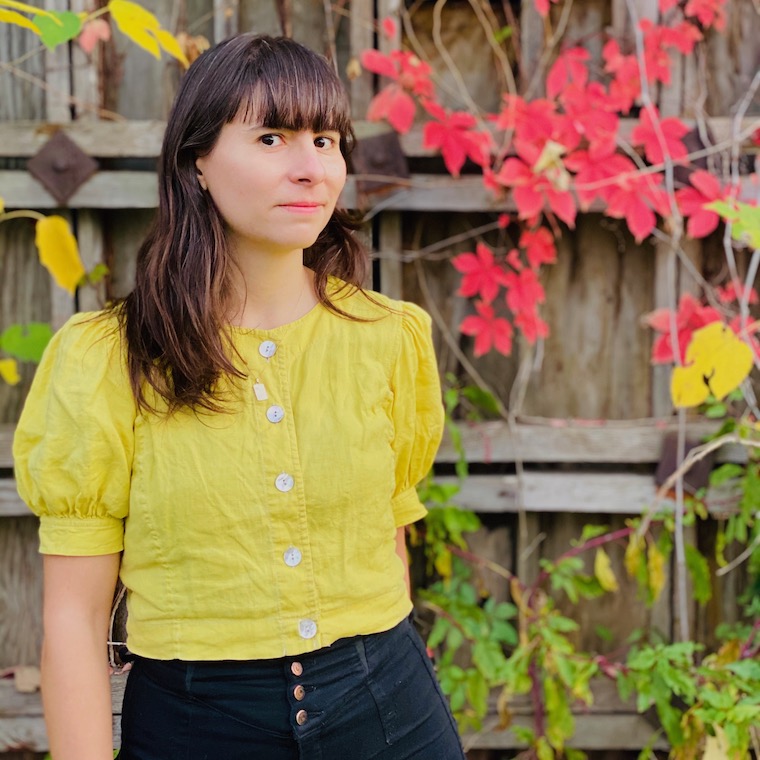 Woman with light skin and long brown hair wearing a yellow blouse and black pants stands in front of a wooden wall covered in colorful fall foliage