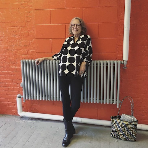 A white woman with gray hair wearing a black and white shirt and black pants leans against a radiator on an orange brick wall
