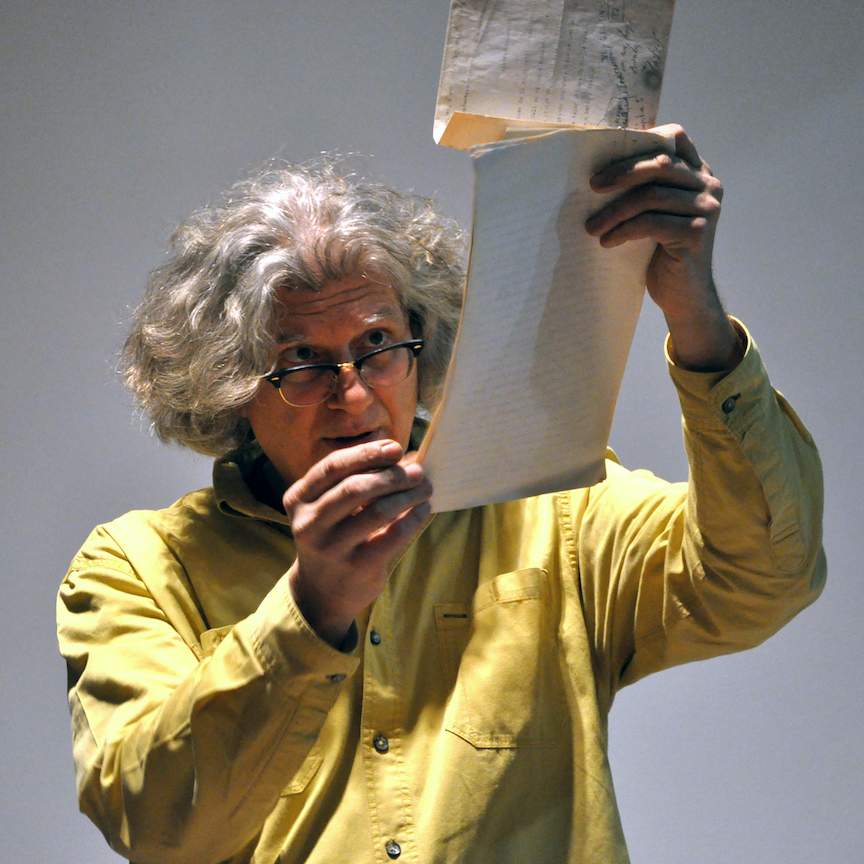 White haired man in yellow shirt wearing glasses holds a manuscript up to the light while reading from it