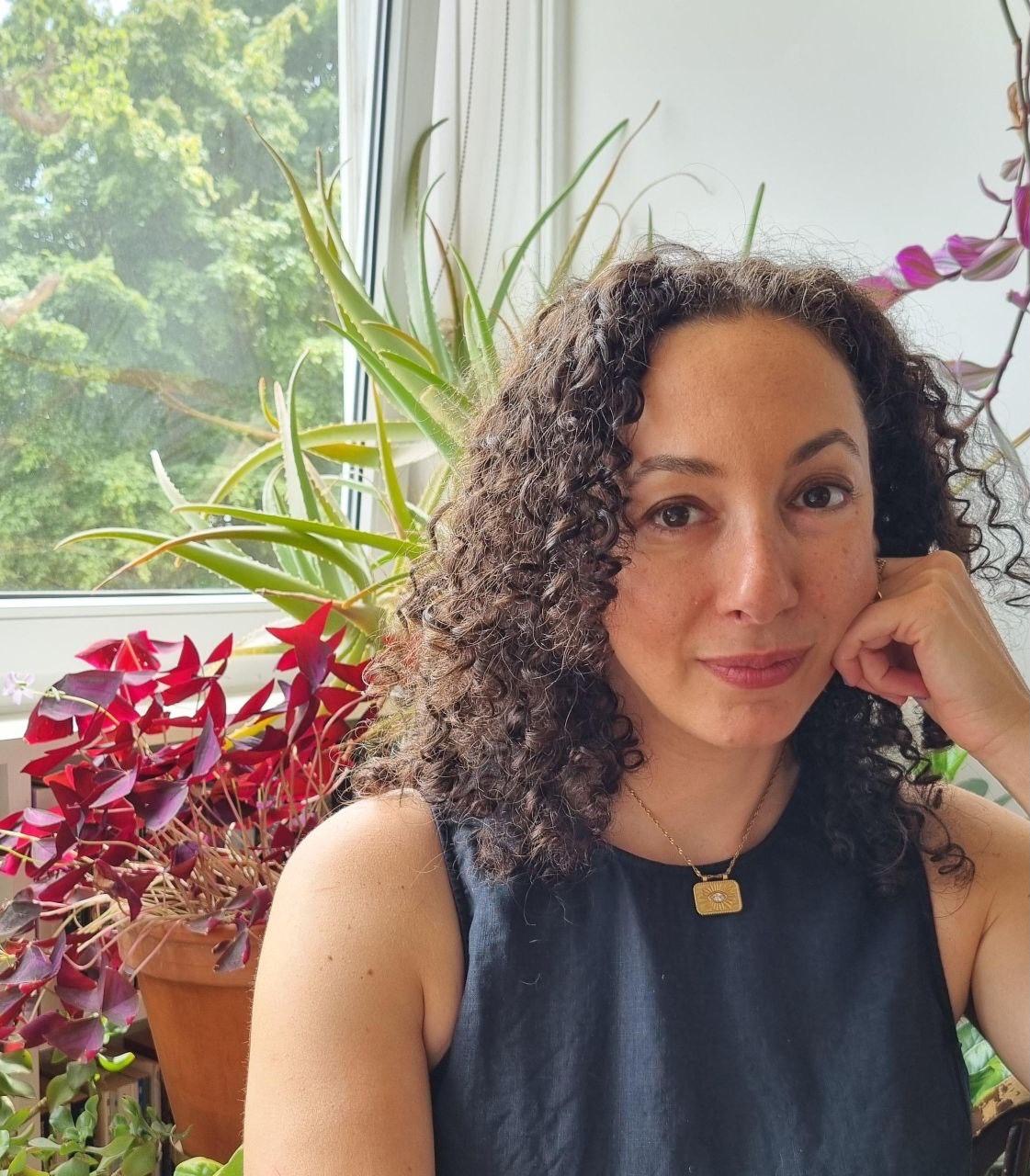 A woman with curly dark hair sits in front of a window with house plants on the sill