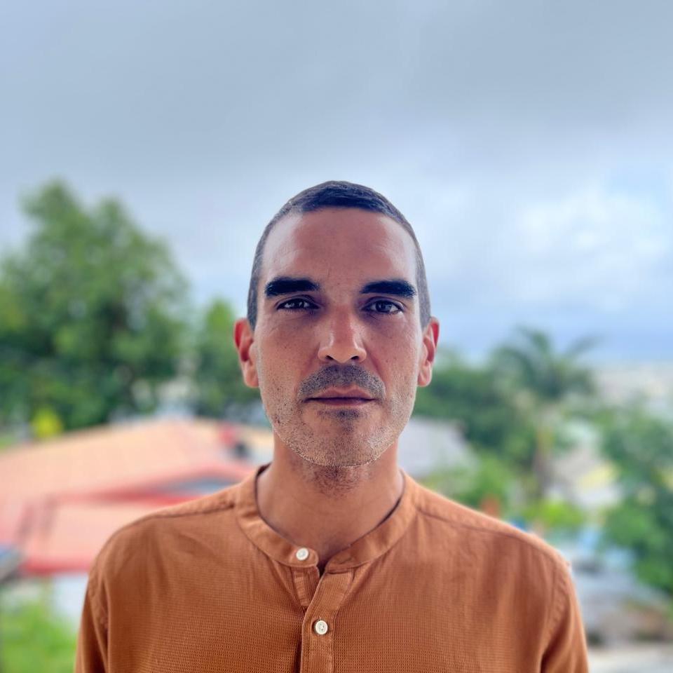 headshot of man with short dark hair in burnt orange shirt standing outside in an equatorial landscape